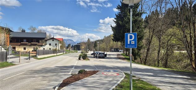 Eine ruhige Straße mit Parkplätzen und einigen Häusern auf der linken Seite. Im Hintergrund sind Bäume und Berge zu sehen, und der Himmel ist klar und blau.