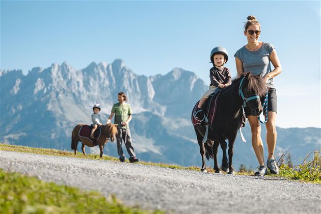 Eine Frau führt ein Pony mit einem kleinen Kind auf dem Rücken. Im Hintergrund sind Berge und eine zweite Familie zu sehen.
