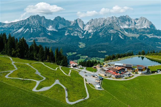 Eine malerische Landschaft mit grünen Wiesen und majestätischen Bergen im Hintergrund. Im Vordergrund liegt eine kleine Siedlung mit einem Teich und gewundenen Wegen.