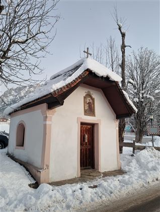Eine kleine Kapelle mit einem Schnee bedeckten Dach. Umgeben von winterlicher Landschaft und schneebedeckten Bäumen.