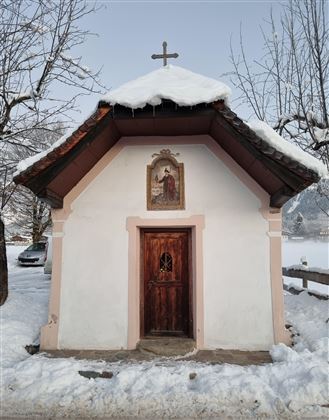 Eine kleine Kapelle mit einem schneebedeckten Dach steht in einer winterlichen Landschaft. Umgeben von schneebedeckten Bäumen und einem ruhigen, friedlichen Ambiente.