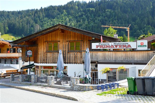 A cozy pizzeria with a wooden facade and outdoor terrace. Surrounded by green mountains and a quiet street.