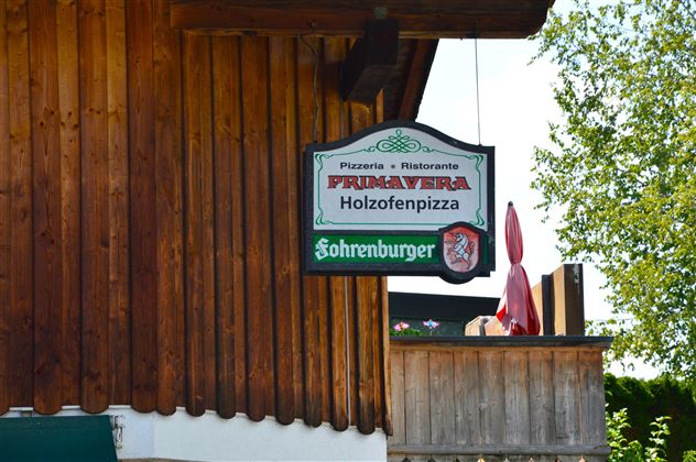 A restaurant sign with the name "Primavera" for wood-fired pizza. The background features wooden cladding and green trees.