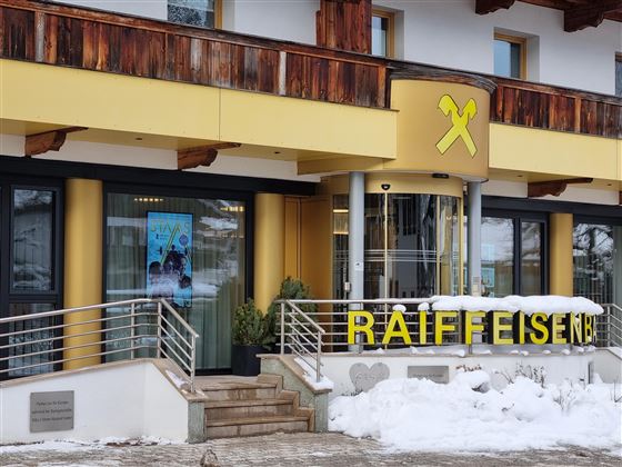 Een modern gebouw van de Raiffeisenbank met een geel bord. Voor de ingang ligt sneeuw, en er is een reclamebord aan de muur.