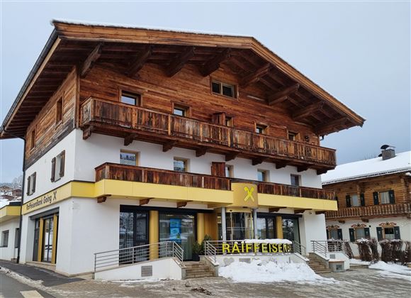 Een traditioneel houten huis met meerdere balkons in de Alpen. Beneden bevindt zich een winkel met een geel bord.