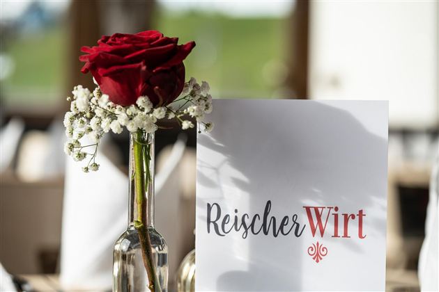 Een elegante tafeldecoratie met een rode roos in een glazen vaas en een menukaart. Op de achtergrond is een groene landschap te zien.