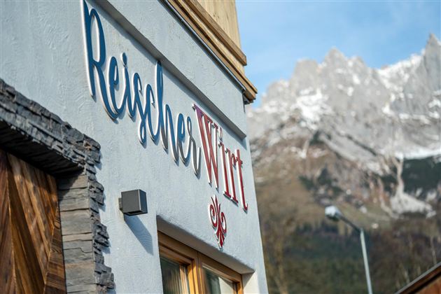 Een gebouw met de tekst "ReiseWirt" in een schilderachtige berglandschap. De omgeving is bergachtig en toont besneeuwde toppen op de achtergrond.