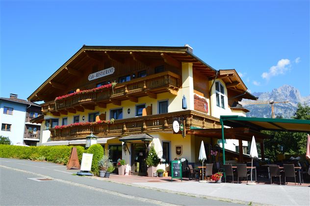A charming alpine hotel with wooden decor and balconies. In the background, beautiful mountains and a clear blue sky can be seen.