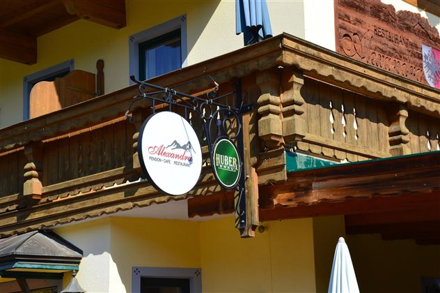 A traditional building with a wooden balcony. In front hang two signs, one with the name "Alpenblick" and the other with a reference to beer.