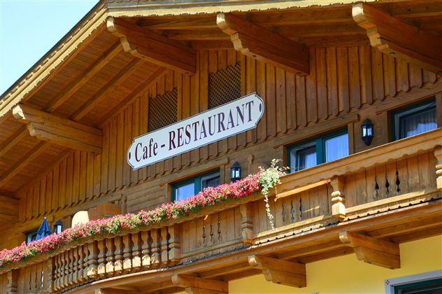 A traditional wooden building with a sign that shows "Cafe - RESTAURANT". Colorful flowers are blooming on the balcony and the windows are small and inviting.
