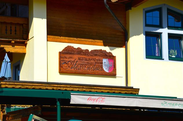 A restaurant named "Alexandra" is indicated by a wooden sign. In the background, parts of a yellow building and windows can be seen.