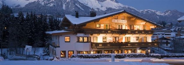 Ein gemütliches Hotel im Schnee mit beleuchteten Fenstern. Im Hintergrund sind schneebedeckte Berge und Bäume zu sehen.