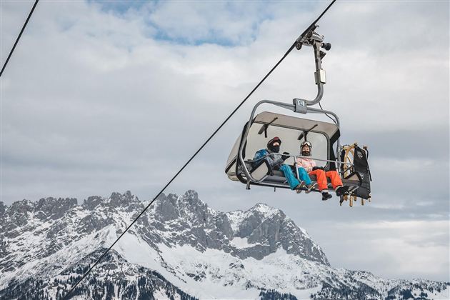 Eine Skiliftkabine mit zwei Fahrgästen schwebt über schneebedeckte Berge. Der Himmel ist bewölkt und die Landschaft wirkt winterlich.
