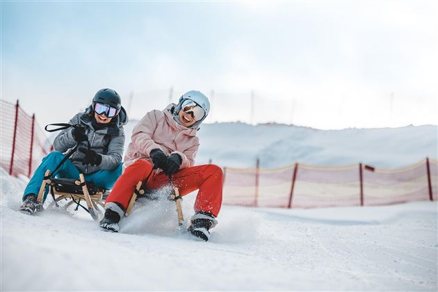 Zwei Personen auf Schlitten im Schnee. Sie haben Spaß und tragen Schutzhelme.