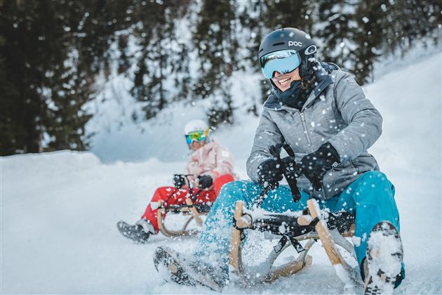 Zwei Personen sitzen auf Schlitten und fahren einen verschneiten Hang hinunter. Schnee fällt und die Umgebung ist von Tannenbäumen umgeben.