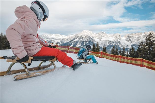 Zwei Kinder rodeln auf einem verschneiten Hang. Im Hintergrund sind schneebedeckte Berge und ein klarer Himmel zu sehen.