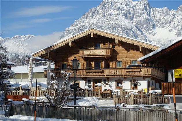 Ein traditionelles, holzverkleidetes Chalet in einer verschneiten Berglandschaft. Im Hintergrund sind beeindruckende Berge und ein klarer blauer Himmel zu sehen.
