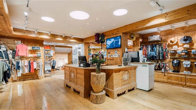 Een gezellig winkeltje met houten decor en een verscheidenheid aan kledingstukken. Bij de kassa staat een plant in een decoratieve pot.