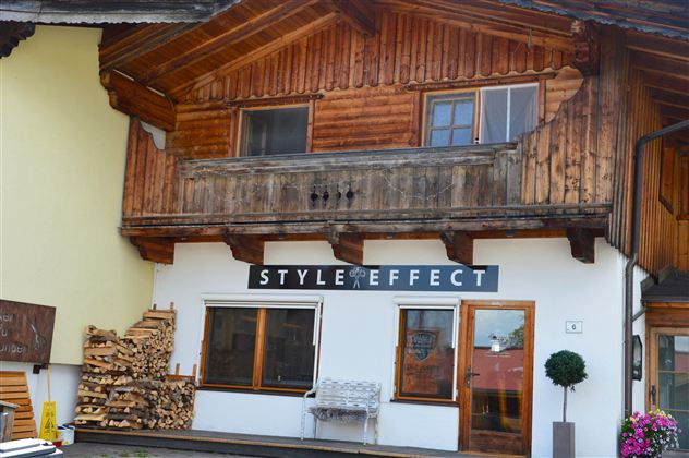 A traditional wooden house with a modern sign "STYLE EFFECT" above the front door. In the foreground, there are wood piles and a small pot with a plant.