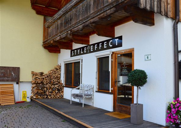 A stylish entrance with the sign "STYLE EFFECT". Next to the door, there is a small table and a plant, while piles of wood can be seen in the background.