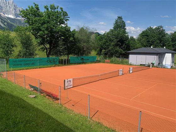Ein Tennisplatz mit roter Asche, umgeben von Bäumen und einem klaren blauen Himmel. Im Hintergrund steht ein kleines Gebäude.