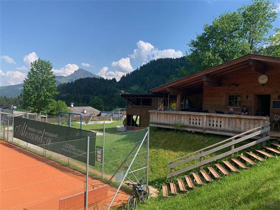 Ein sportlicher Bereich mit einem Tennisplatz und einem Holzhaus im Hintergrund. Die Umgebung ist grün, umgeben von Hügeln und Bäumen, und der Himmel ist blau mit einigen Wolken.