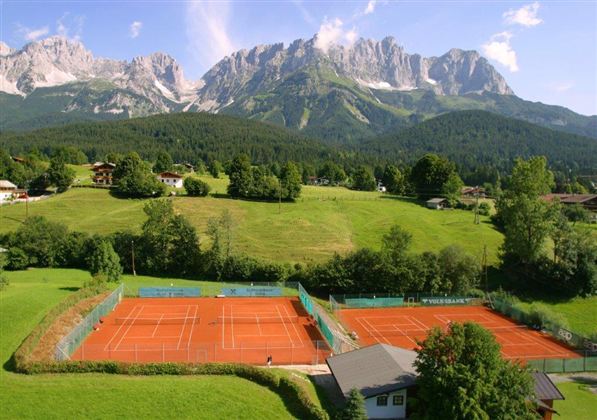 Ein schöner Tennisplatz mit roten Sandbelag in einer grünen Landschaft. Im Hintergrund erheben sich beeindruckende Berge unter einem blauen Himmel.