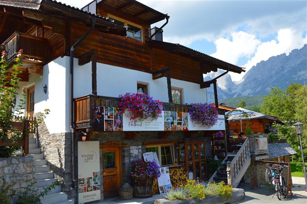 A charming wooden house with a balcony full of flowers. In the background, there are mountains and a clear sky.