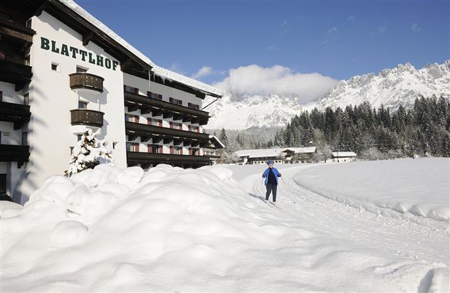 Ein Hotelgebäude mit dem Namen "Blatthof" liegt in einer schneebedeckten Landschaft. Im Hintergrund sind schneebedeckte Berge und ein klarer blauer Himmel zu sehen.