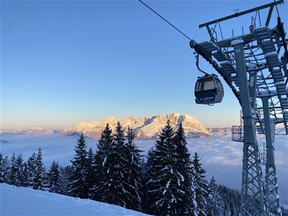 Een kabelbaan rijdt over besneeuwde bomen in de bergen. Op de achtergrond zijn hoge bergen onder een heldere lucht te zien.