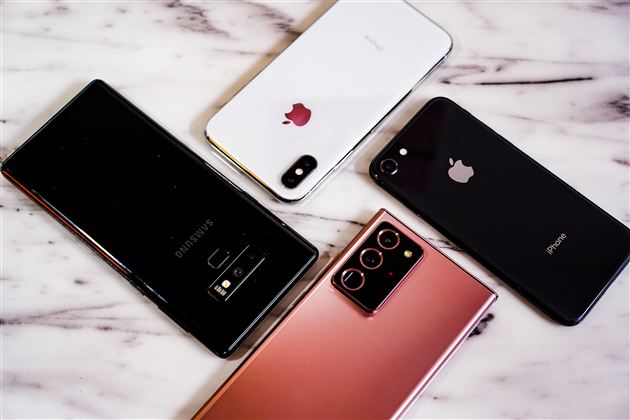 Four smartphones are lying on a marble surface. Two devices are from Apple and two are from Samsung.