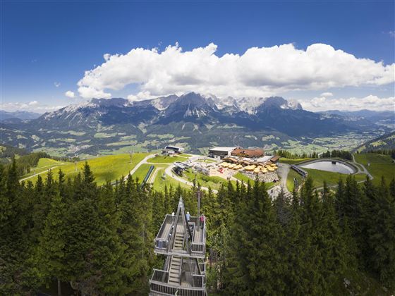 An impressive mountain landscape with lots of greenery and high peaks.  
In the foreground, there is a mountain station with cozy seating areas and paths visible.