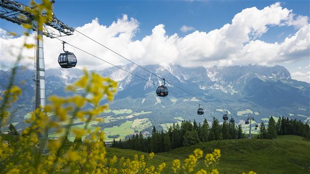 A landscape with mountain gondolas traveling over green meadows. In the background, impressive mountains and a blue sky with clouds can be seen.