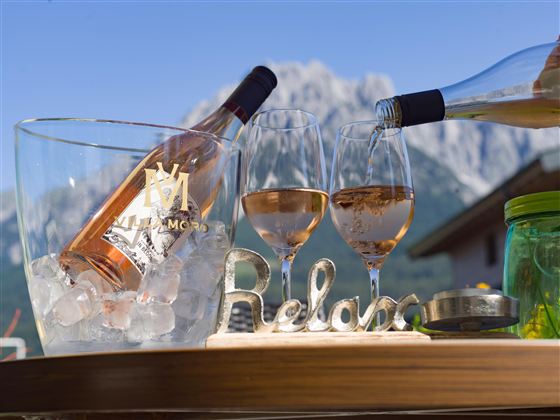 Ein elegantes Getränkeset mit einer Flasche Roséwein und zwei Gläsern steht auf einem Holztisch. Im Hintergrund sind Berge und ein klarer Himmel zu sehen.