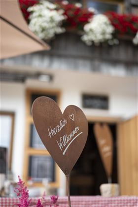 Een hartvormig bord met de inscriptie "Hartelijk Welkom" staat op een tafel. Op de achtergrond zijn bloeiende planten en een gezellige sfeer te zien.
