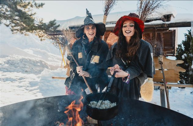 Twee vrouwen in creatieve heksen kostuums staan bij het vuur en koken in een grote pot. Op de achtergrond is sneeuw te zien, wat een winterse sfeer creëert.