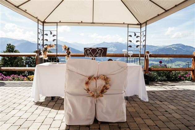A beautifully decorated wedding table under a tent with a picturesque mountain landscape in the background. The chairs are adorned with white covers and a heart-shaped wreath.