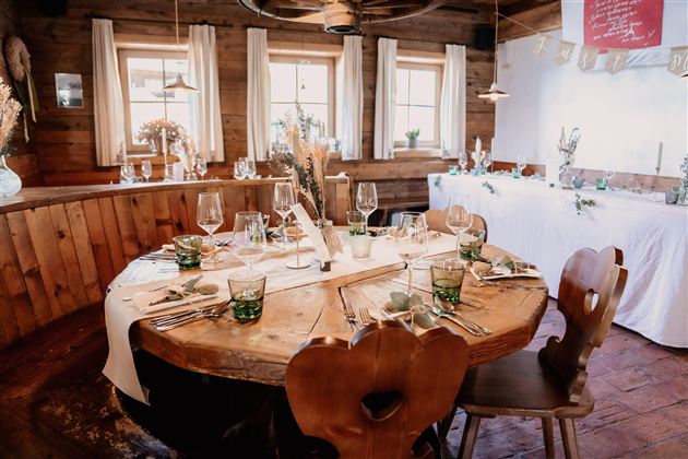 A rustic dining area with a round wooden table, lovingly set with glasses and cutlery. The walls are made of wood and there are large windows with soft light.