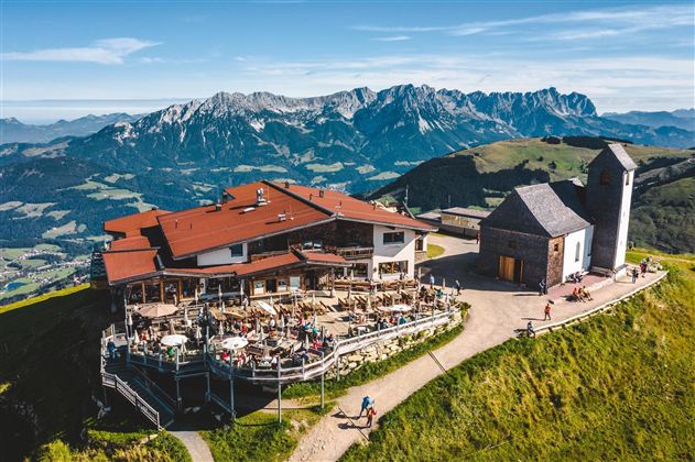Een berghut met buitenterras en uitzicht op de bergen. Helderblauwe lucht en groene weilanden omringen de hut.