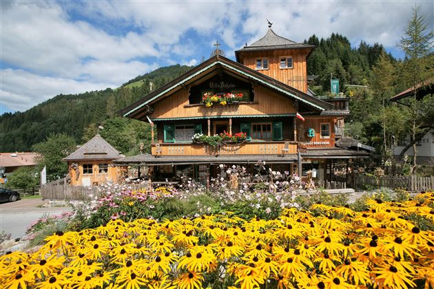 Ein charmantes, holzverkleidetes Gebäude mit bunten Blumen im Vordergrund. Im Hintergrund sind grüne Hügel und ein blauer Himmel zu sehen.