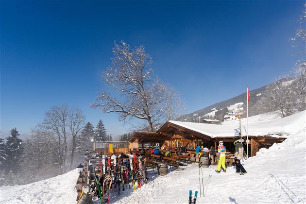 Eine alpine Skihütte steht im Schnee unter einem blauen Himmel. Skifahrer genießen die winterliche Landschaft und machen eine Pause.