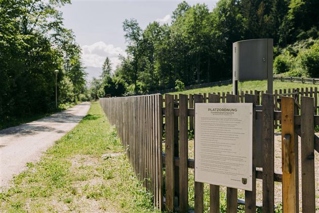 A gravel path leads through a green landscape with trees. An information sign stands at a wooden fence along the path.