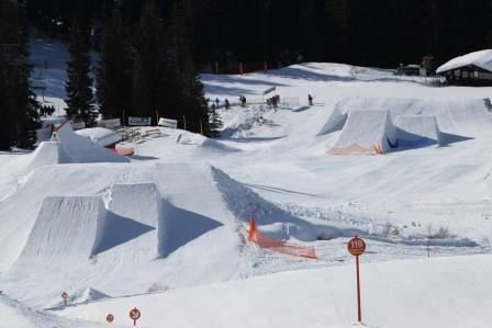 Eine verschneite Landschaft mit mehreren Sprüngen für Wintersportler. Im Hintergrund sind Bäume und ein Gebäude sichtbar.