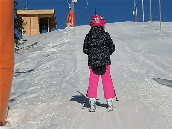 A child in pink ski clothing stands on a snowy slope. In the background, a ski lift station and blue sky are visible.