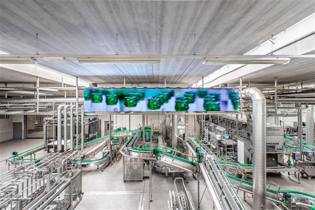 Een moderne productie-installatie met complexe pijpleidingen en transportbanden. Boven de machines hangt een grote monitor.