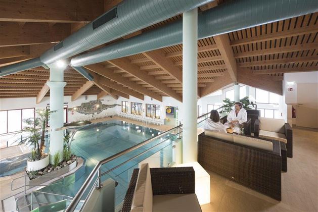 Ein eleganter Innenpool mit Holzdecke und Pflanzen. Im Vordergrund sitzen zwei Personen auf einer gemütlichen Sitzgruppe.