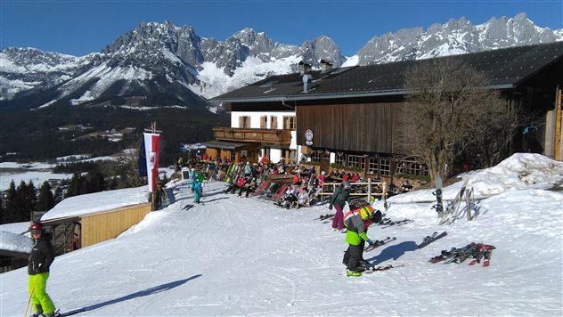 Een alpine skigebied met besneeuwde bergen en een houten huis op de achtergrond. Meerdere skiërs en snowboarders genieten van de piste.