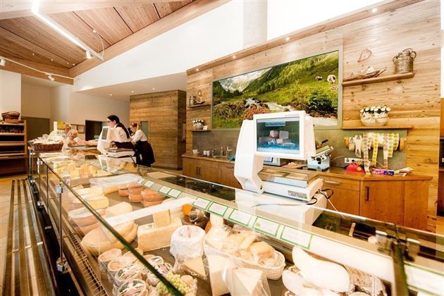 Een gezellige kaaswinkel met een selectie van verschillende kaassoorten. Achtergrondmedewerkers werken aan de toonbank en decoratieve elementen creëren een uitnodigende sfeer.