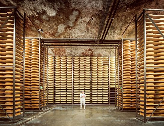 Een grote opslaghal met veel kazen, netjes gestapeld op planken. Een persoon staat in het midden van de hal.