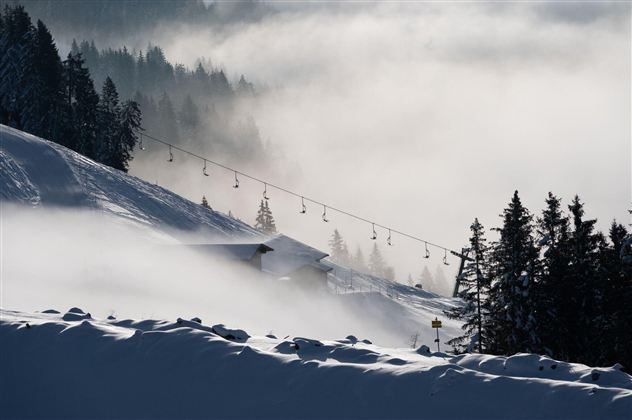 A winter landscape with fresh snow and fog. Ski lifts are visible in the background, surrounded by snow-covered trees.